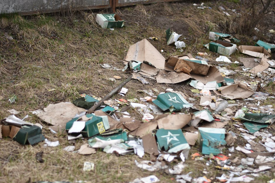 An outdoor scene showing a patch of uneven grassy ground with scattered debris, including crumpled and torn cardboard boxes, some with printed logos in shades of green and white, and broken fragments of paper and plastic. The cardboard appears weathered, with some flattened or torn into irregular shapes, while the paper debris is crumpled and mixed among the boxes. In the background, there is a wooden fence or barrier, partially obscured by the debris, and a few sparse patches of dry, brown vegetation. The scene is illuminated by natural daylight, creating a dull, somewhat overcast atmosphere. This image depicts unmanaged waste, consistent with private rubbish disposal or alternative collection scenarios, highlighting the importance of professional rubbish removal services such as those offered by Rubbish Removal Earls Court in managing and clearing such litter from outdoor areas.