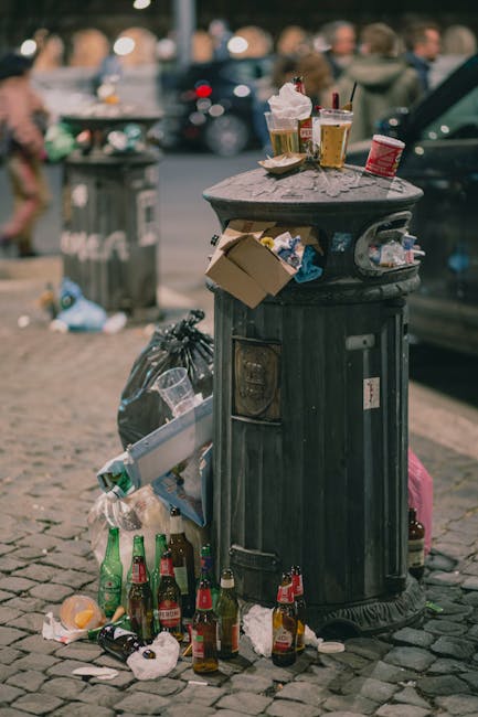 A waste collection worker, wearing a bright red and yellow uniform, is standing at the rear of a large red rubbish collection truck on a roadside. The worker is in the process of emptying a black plastic bag filled with waste into the truck's open hopper. The truck is parked partly on a curb, adjacent to a paved road with visible lane markings. Behind the worker, there are small scattered debris and a black motorcycle parked nearby with a yellow and red striped safety sign attached to its rear rack. The scene is set in an outdoor urban environment with trees and utility poles in the background, and overcast weather creating diffused natural lighting. The image reflects a routine waste collection operation, which may belong to a private rubbish removal service such as Rubbish Removal Earls Court, highlighting alternative waste handling methods outside of local authority disposal systems.