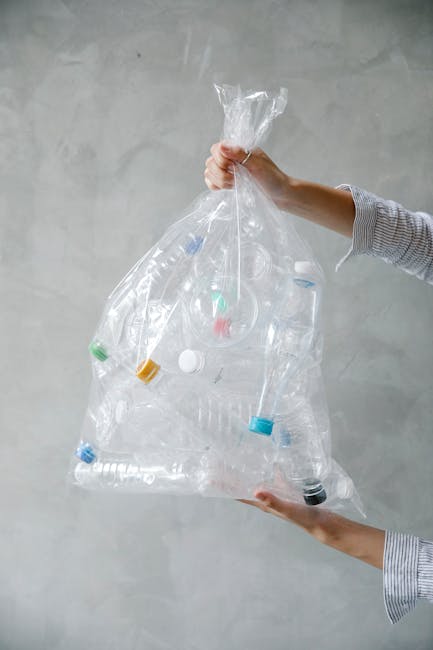 A person wearing a light-colored, long-sleeved shirt is holding a large, transparent plastic bag filled with various used plastic bottles and containers. The bag is tied at the top and tilted slightly downward, with the person's right hand gripping the gathered neck of the bag and the left supporting the bottom. The plastic bottles inside the bag vary in size and shape, with some having coloured caps in green, yellow, blue, and black, and some with labels or markings visible through the clear plastic. The background is a neutral, smooth, light grey wall with a subtle texture, providing a plain backdrop that draws focus to the bag and its contents. This scene subtly reflects the practice of independent plastic waste collection, which can be associated with private rubbish removal services such as those offered by Rubbish Removal Earls Court.