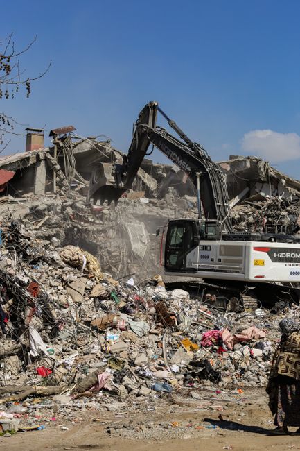 A large hydraulic excavator with a black arm and grey body is actively engaged in demolishing a building, with parts of concrete, bricks, and debris falling as the structure is partially collapsed. The scene shows a mound of mixed rubble, including broken concrete slabs, twisted rebar, and various discarded materials such as plastics and textiles, scattered across the foreground. The excavator's cab is enclosed with glass and positioned to the right, while the demolished building remnants extend across the background under a bright blue sky with a few white clouds. In the lower right corner, a person wearing a patterned garment is partially visible, observing the scene. The setting appears to be an outdoor construction or demolition site, consistent with the activity of clearing waste materials for disposal or recycling, a process aligned with private rubbish removal services like those offered by Rubbish Removal Earls Court. The natural daylight highlights the textures of the rubble and machinery, emphasizing the scale of the demolition work involved in waste clearance operations.